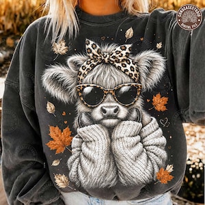 May include: A charcoal gray sweatshirt featuring a graphic of a fluffy Highland cow wearing a leopard print bow and black sunglasses. The cow is depicted in a thick knit sweater, surrounded by scattered orange and brown autumn maple leaves.
