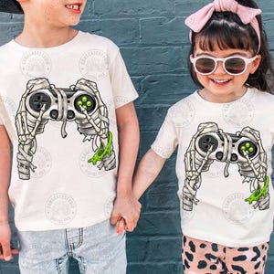 Op de afbeelding: Twee off-white t-shirts met een grafische print van een videogamecontroller gewikkeld in mummiebandages. De controller wordt vastgehouden door skeletachtige handen, met felgroen slijm dat van één kant van de controller druipt.