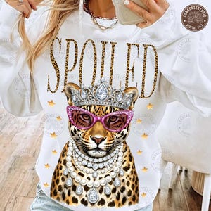 May include: White hoodie with the word "SPOILED" in a leopard print font. The graphic features a leopard wearing a jeweled crown, pink sunglasses, and a pearl necklace. Gold stars are scattered around the design.