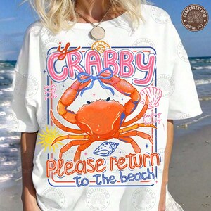 Pode incluir: Camiseta branca com uma ilustração de caranguejo laranja com um laço azul. O texto diz "If Crabby, Please Return to the Beach!" em rosa e vermelho. Uma camiseta gráfica divertida com tema de praia.
