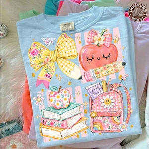 Könnte beinhalten: Ein hellblaues T-Shirt mit einem Grafikdruck, der eine große gelbe Vichy-Schleife, einen lächelnden Apfel mit Bleistift, einen Bücherstapel mit einem Blumenapfel darauf und einen rosa Rucksack mit Gänseblümchen zeigt, akzentuiert durch kleine goldene Sterne.
