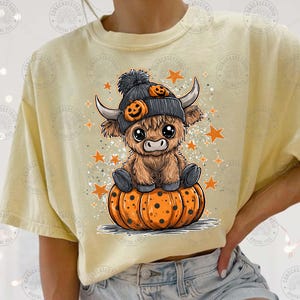 May include: A light yellow t-shirt featuring a cartoon Highland cow wearing a black hat with pumpkin accents, sitting on an orange pumpkin. Orange stars and glitter surround the cow, creating a Halloween theme.