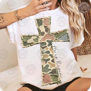 Op de afbeelding: Wit T-shirt met een groot kruisontwerp gemaakt van camouflage stoffen patches. Het kruis heeft een gerafelde rand. De woorden "He is Risen" staan verticaal aan de rechterkant van het kruis in een handschriftstijl.