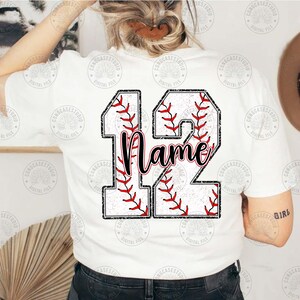 May include: White t-shirt with a baseball-themed design featuring the number 12. The number is made up of two baseballs with red stitching and a white background. The word "Name" is written in black cursive lettering inside the number.