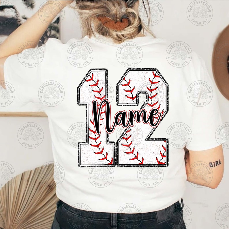May include: White t-shirt with a baseball-themed design featuring the number 12. The number is made up of two baseballs with red stitching and a white background. The word "Name" is written in black cursive lettering inside the number.