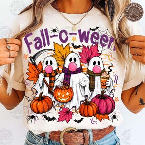 May include: A cream-colored t-shirt featuring a graphic of three white ghosts wearing scarves and blowing pink bubblegum. The ghosts are surrounded by orange and pink pumpkins, autumn leaves, bats, spiders, and the purple text Fall-c-ween.