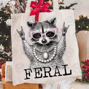 May include: A cream-colored tote bag featuring a raccoon wearing pink sunglasses and a pearl necklace, with the word "FERAL" in bold black letters. The raccoon is making a rock-and-roll hand gesture.