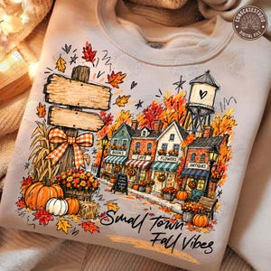 May include: A beige sweatshirt featuring a graphic print of a small town street with shops labelled Books, Cafe, Flowers, and Antiques. The design includes a wooden signpost, a water tower, pumpkins, and autumn leaves with the text Small Town Fall Vibes.