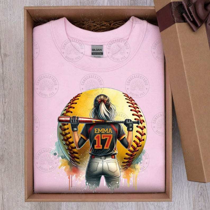 Peut inclure: T-shirt rose avec un motif repr&eacute;sentant une joueuse de softball tenant une batte devant un grand ballon de softball jaune avec des coutures rouges. La joueuse porte un maillot noir avec le nom "Emma" et le num&eacute;ro "17" au dos. Le motif a un effet aquarelle.