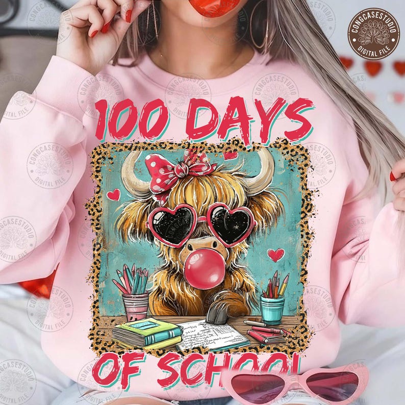 May include: A pale pink sweatshirt with the text "100 DAYS OF SCHOOL" in red and a leopard print border. The graphic features a cartoon Highland cow wearing heart-shaped sunglasses and blowing a bubble. The cow is at a desk with pencils and books.