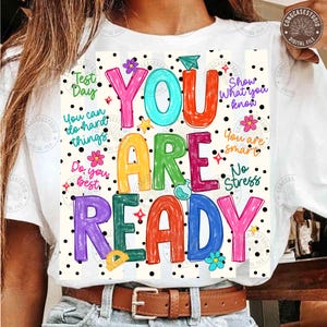Può includere: T-shirt bianca con una grafica colorata che recita "YOU ARE READY". Il design presenta le parole in vari colori vivaci, accentate da fiori, stelle e pois. Il testo aggiuntivo include "Test Day" e "You can do hard things."