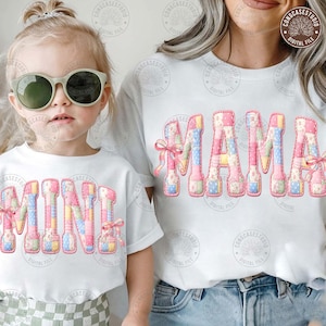 May include: White t-shirts with the words "MAMA" and "MINI" in a patchwork quilt design. The letters are in pink, blue, and yellow with pink bows. The shirts are worn by a mother and child.