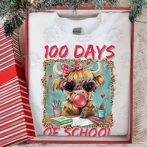 May include: White sweatshirt featuring a Highland cow graphic with heart-shaped sunglasses and a bubble. The text "100 Days of School" is in red. The sweatshirt is in a red gift box. The image is in a red gift box.