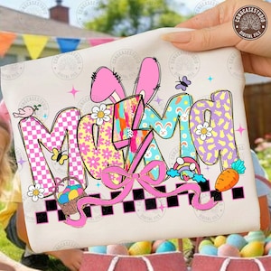 May include: A cream-colored graphic tee with the word "MOM" in colorful, patterned letters. The design includes bunny ears, flowers, a basket of Easter eggs, a rainbow, and a carrot. The tee is held up by a hand.