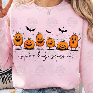 May include: A light pink crewneck sweatshirt featuring a graphic of six orange jack-o'-lanterns with various facial expressions, black bats, and small stars. The text "spooky season" is printed in a black cursive font below the pumpkins.