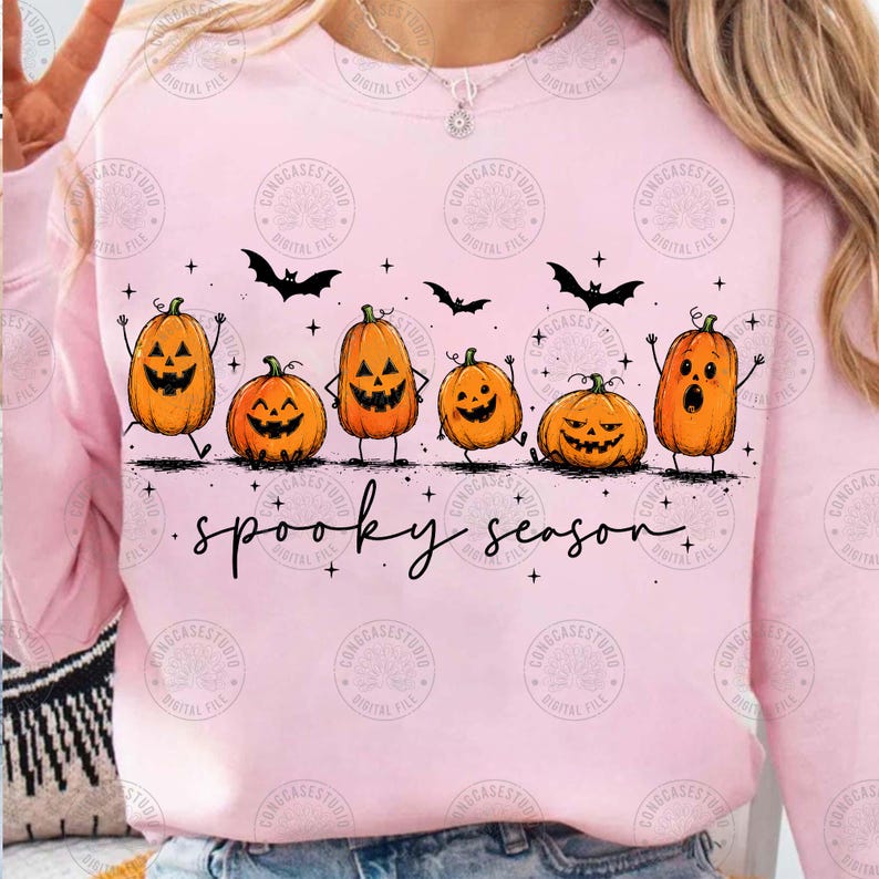 May include: A light pink crewneck sweatshirt featuring a graphic of six orange jack-o'-lanterns with various facial expressions, black bats, and small stars. The text "spooky season" is printed in a black cursive font below the pumpkins.
