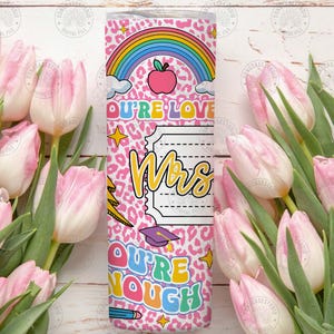 Peut inclure: Un gobelet rose avec un motif l&eacute;opard, un arc-en-ciel et le texte "OU'RE LOVE Mrs OU'RE NOUGH". Le gobelet pr&eacute;sente &eacute;galement une pomme, des &eacute;toiles, un crayon et un chapeau de remise de dipl&ocirc;me. Le gobelet est entour&eacute; de tulipes roses.