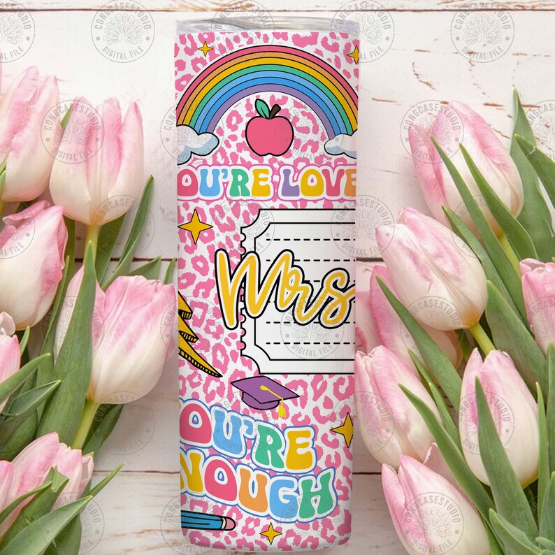 Peut inclure: Un gobelet rose avec un motif l&eacute;opard, un arc-en-ciel et le texte "OU'RE LOVE Mrs OU'RE NOUGH". Le gobelet pr&eacute;sente &eacute;galement une pomme, des &eacute;toiles, un crayon et un chapeau de remise de dipl&ocirc;me. Le gobelet est entour&eacute; de tulipes roses.
