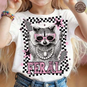 May include: White t-shirt featuring a raccoon wearing pink sunglasses and a pearl necklace. The raccoon is surrounded by pink stars, black and white checkered patterns, and the word "FERAL" in pink letters.