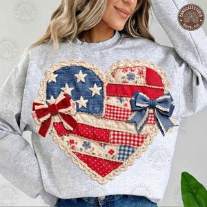 Könnte beinhalten: Ein graues Sweatshirt mit einem herzförmigen Design in patriotischen Farben. Das Herz besteht aus Denim, rot-weiß gemusterten Stoffen, weißen Sternen und zwei dekorativen Schleifen. Das Design ist mit einer cremefarbenen Spitzenborte umrandet.