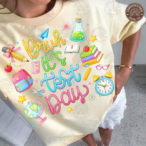 Peut inclure: Un t-shirt jaune clair avec un motif graphique coloré sur le thème de l'école. Le motif comprend le texte "Bruh it's Test Day" avec des illustrations d'un crayon, de livres, d'un sac à dos et d'un réveil.