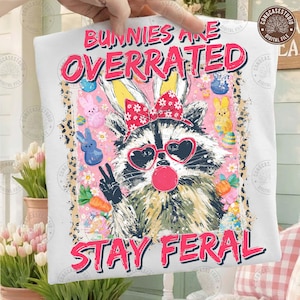 May include: White t-shirt with a raccoon graphic wearing heart-shaped sunglasses and a bow, blowing a bubblegum bubble. The text reads "Bunnies Are Overrated, Stay Feral". Easter-themed design with colorful accents.