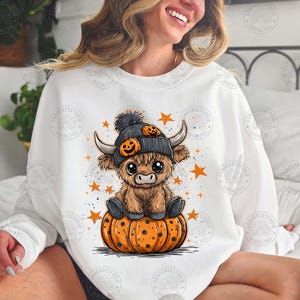 May include: A white sweatshirt featuring a cartoon Highland cow wearing a black hat with pumpkin accents, sitting on an orange pumpkin. Orange stars are scattered around the cow. The design is a digital file.