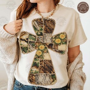 May include: A cream-coloured t-shirt with a cross design. The cross is a patchwork of camouflage, tree bark, and floral patterns, with yellow roses. The design is outlined with a stitched border.