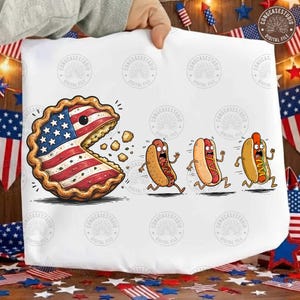 May include: A white graphic print featuring a large, anthropomorphic pie decorated with the American flag pattern, appearing to chase three cartoon hot dogs. The pie has a wide, open mouth and is shown eating crumbs as the hot dogs run away.