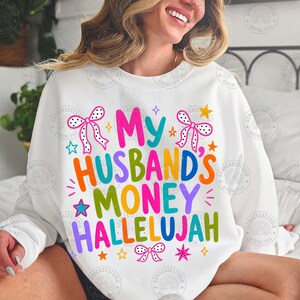 K&ouml;nnte beinhalten: Ein wei&szlig;es Sweatshirt mit Rundhalsausschnitt und dem bunten Schriftzug My HUSBAND'S MONEY HALLELUJAH in einer verspielten Schriftart. Das Design ist mit verschiedenen kleinen Sternen und drei gepunkteten Schleifen in Pink und Schwarz verziert.