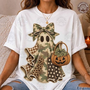 May include: A white t-shirt featuring a central graphic of a ghost wearing a large bow. The ghost has a camouflage pattern with sections of leopard print, polka dots, and checkered fabric. It holds an orange jack-o'-lantern bucket with green leaves.