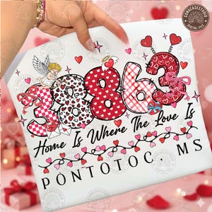 May include: White t-shirt featuring the phrase "Home Is Where The Love Is" with the number "3863" in a heart-themed design. The design includes hearts, bows, and an angel, perfect for Valentine's Day or a romantic gift.