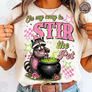 May include: A cream-colored t-shirt featuring a graphic of a raccoon wearing a pink apron and stirring a black cauldron filled with green liquid. The text reads On my way to STIR the Pot, surrounded by pink and green stars and checkered patterns.