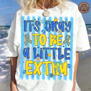 May include: A white t-shirt featuring a blue and yellow graphic with the text IT'S OKAY TO BE A LITTLE EXTRA. The design includes vertical light blue stripes, yellow and blue puzzle piece ribbons, and small flower icons throughout the print.