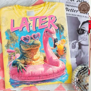 May include: Yellow t-shirt with a graphic featuring an alligator wearing heart-shaped sunglasses and a pink flamingo float. The alligator is in a pink floatie, holding a drink. The word "LATER" is printed above the alligator in pink letters.