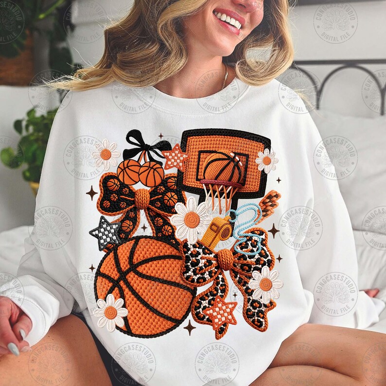 May include: White sweatshirt featuring a basketball design. The design incorporates basketballs, a hoop, bows, stars, and flowers in orange, black, and white. The sweatshirt has long sleeves and a crew neck.