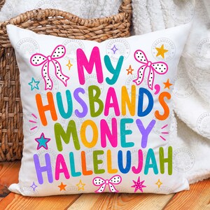 K&ouml;nnte beinhalten: Ein wei&szlig;es quadratisches Dekokissen mit dem bunten Schriftzug "MY HUSBAND'S MONEY HALLELUJAH" in verschiedenen leuchtenden Farben. Das Design ist mit rosa-wei&szlig; gepunkteten Schleifen und kleinen bunten Sternen auf der Oberfl&auml;che verziert.