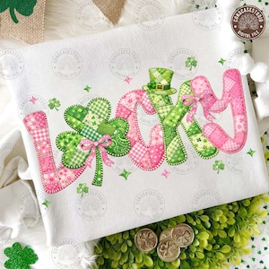 Może przedstawiać: Biała bluza z napisem "LUCKY" w literach patchwork. Litery są różowe i zielone z akcentami koniczyny i kokardek. Zielony cylinder ze złotą klamrą znajduje się nad literą "Y".