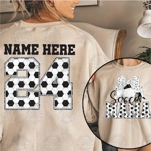 K&ouml;nnte beinhalten: Ein beiger Pullover mit einem schwarz-wei&szlig;en Fu&szlig;ball-Design. Der Text "NAME HERE" und die Nummer "24" befinden sich auf der R&uuml;ckseite des Pullovers. Ein zweiter beiger Pullover mit einem schwarz-wei&szlig;en Fu&szlig;ball-Design und dem Text "Soccer Mama" ist auf dem Bild zu sehen.