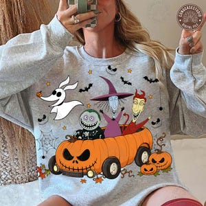 May include: A light gray crewneck sweatshirt featuring a graphic print of characters from The Nightmare Before Christmas riding in a large orange jack-o'-lantern car, accompanied by Zero the ghost dog, bats, spiders, and small pumpkins.