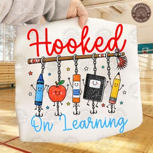 May include: A white graphic print featuring the red text Hooked and blue text On Learning. The design includes a horizontal fishing rod with five hanging school supplies: a blue crayon, a red apple, an orange glue stick, a black notebook, and a yellow pencil, all with smiling faces and fishing hooks.