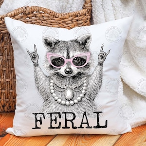 May include: White throw pillow with a black and white illustration of a raccoon wearing pink sunglasses and a pearl necklace. The raccoon is making a rock and roll hand gesture. The word "FERAL" is printed in black below the raccoon.
