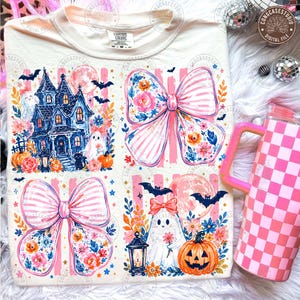 Puede incluir: Una camiseta color crema con un estampado gráfico de dos grandes lazos rosas a rayas con detalles florales, una casa encantada azul oscuro, dos fantasmas blancos, un gato negro, calabazas naranjas, murciélagos y una luna llena. Un vaso de cuadros rosa y blanco está al lado.