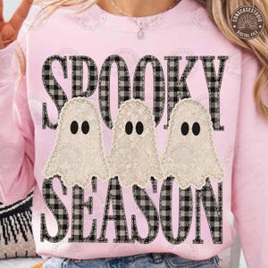 May include: A light pink sweatshirt featuring the text SPOOKY SEASON in a black and white buffalo plaid pattern. Three cream-colored lace ghosts with black oval eyes are positioned between the two words, creating a layered graphic design.