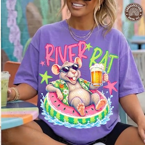 May include: A purple t-shirt featuring a graphic of a rat wearing sunglasses and a floral shirt, floating in a watermelon inner tube while holding a glass of beer. The text "RIVER RAT" is printed above the rat in pink and green lettering.