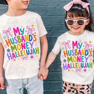 K&ouml;nnte beinhalten: Zwei cremefarbene Kurzarm-T-Shirts mit dem bunten Schriftzug My Husband's Money Hallelujah in Pink, Orange, Gr&uuml;n und Blau. Das Design enth&auml;lt kleine rosa Schleifen, gelbe Sterne und violette Glitzerakzente um die kr&auml;ftige Schrift herum.