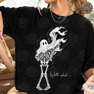 May include: A black t-shirt featuring a white graphic illustration of a skeleton wearing a ghost sheet. The skeleton has its hands crossed over its chest, and the ghost sheet flows upward. The text "Well, shit..." is printed in white script.
