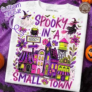 Puede incluir: Una camiseta blanca con un gráfico colorido de Halloween que muestra un pueblo pequeño, una bruja en escoba, un fantasma y un caldero. El texto dice "SPOOKY IN A SMALL TOWN" y "688824" en una señal. El diseño incluye detalles en morado y verde lima.