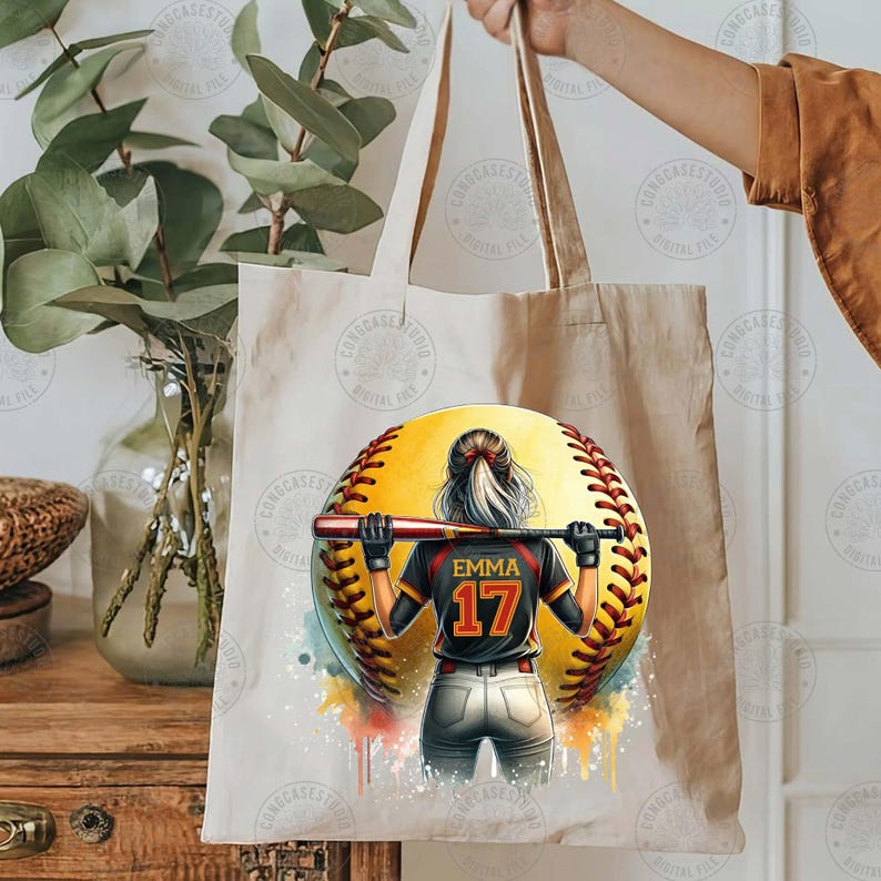 Peut inclure: Un sac fourre-tout blanc avec une illustration color&eacute;e d'une joueuse de softball tenant une batte. La balle de softball est jaune avec des coutures rouges et la joueuse porte un maillot noir avec le nom "Emma" et le num&eacute;ro "17" au dos.