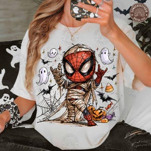 May include: A cream-colored t-shirt featuring a graphic of a Spider-Man character wrapped in mummy bandages, making a rock-and-roll hand gesture. The design includes small white ghosts, black bats, a spiderweb, pumpkins, and candy corn pieces.